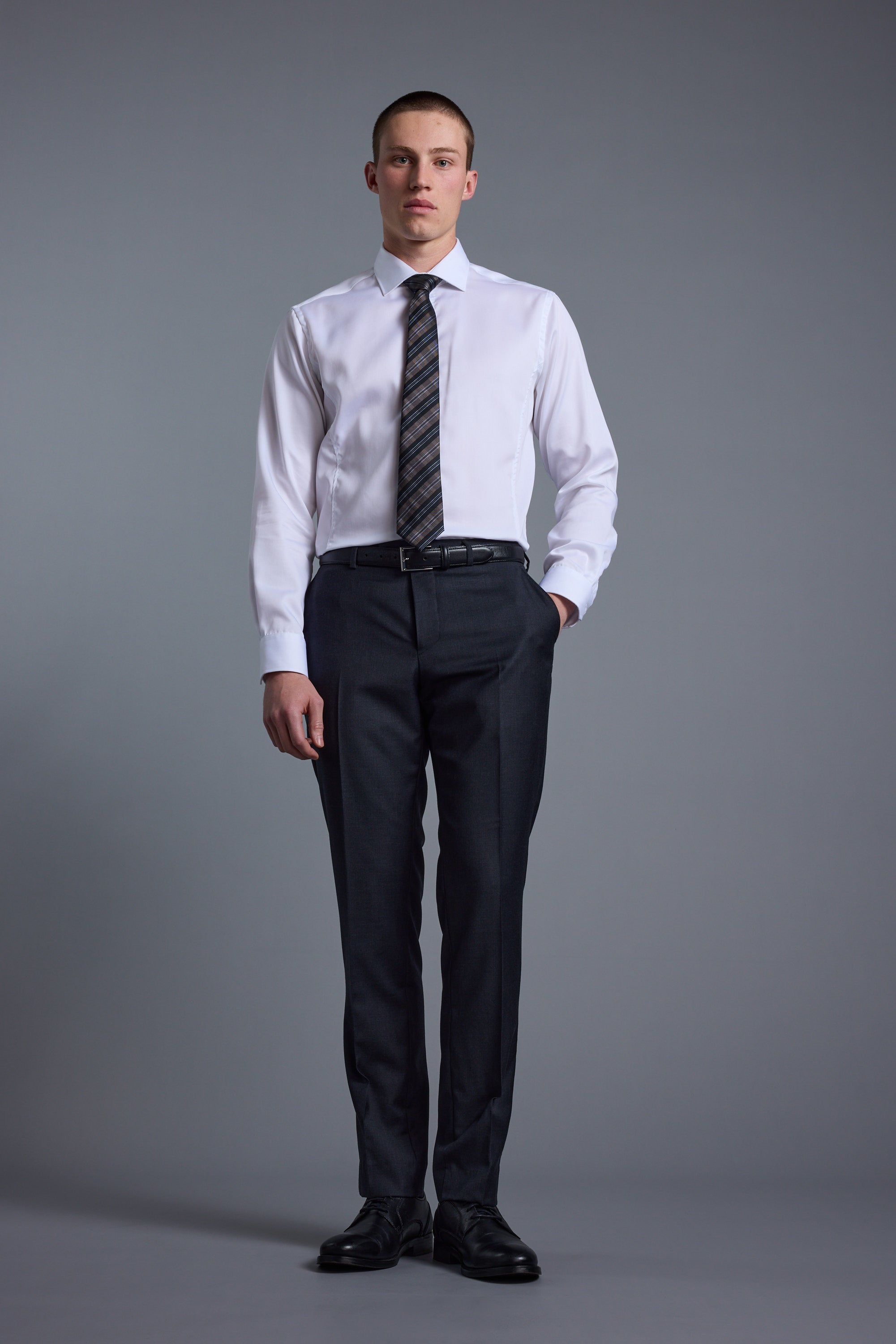 A man stands facing forward against a gray background, wearing Barkers Mayfair ZQrx Suit Trousers paired with a white dress shirt, dark tie, and black dress shoes, with one hand in his pocket.