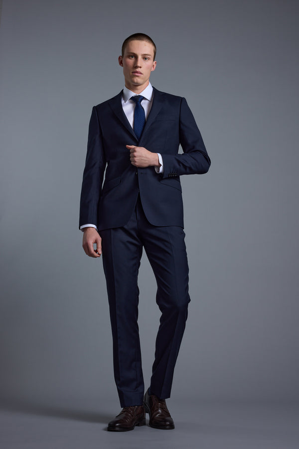 A man stands against a gray background wearing the Barkers Mayfair ZQ Suit Jacket with a white dress shirt, blue tie, and brown shoes. He faces forward, adjusting his suit jacket.