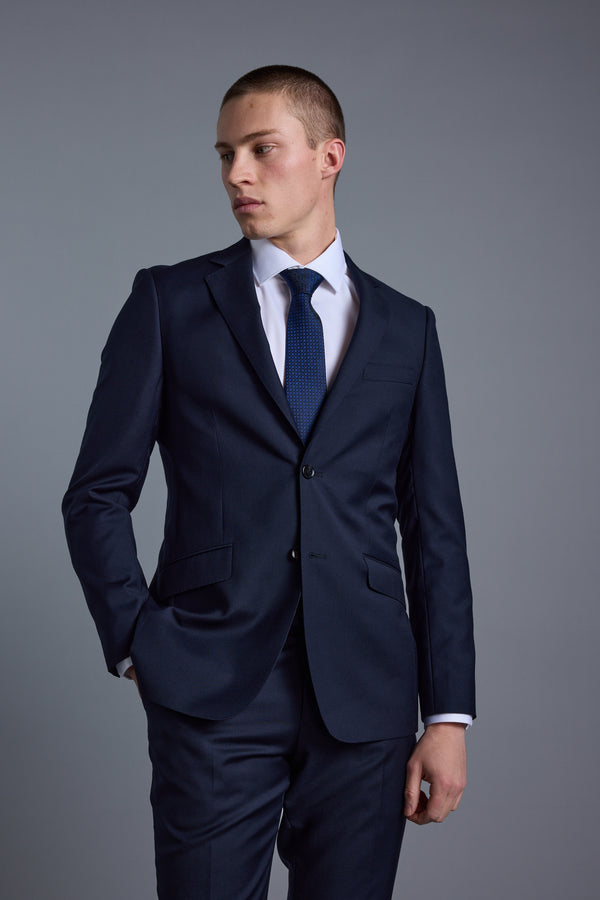 A young man wearing the Barkers Mayfair ZQ Suit Jacket, a white dress shirt, and a blue tie stands against a plain gray background, looking to his left with one hand in his pocket.