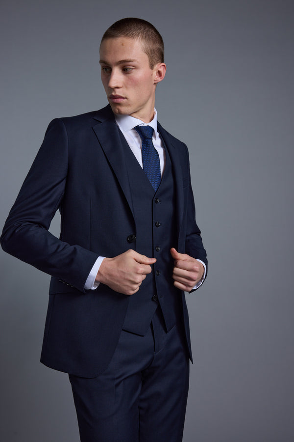 A young man wearing the Barkers Mayfair ZQ Suit Jacket, with a matching vest, white shirt, and blue tie stands against a plain gray background, looking to his left while adjusting his jacket.