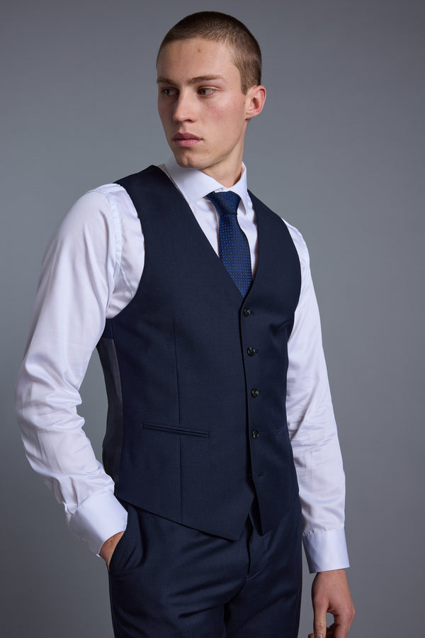 A young man with short hair poses against a gray background wearing the Barkers Mayfair ZQ Waistcoat and matching trousers made from ZQ-certified New Zealand merino wool, with a white dress shirt and blue tie, one hand in his pocket.