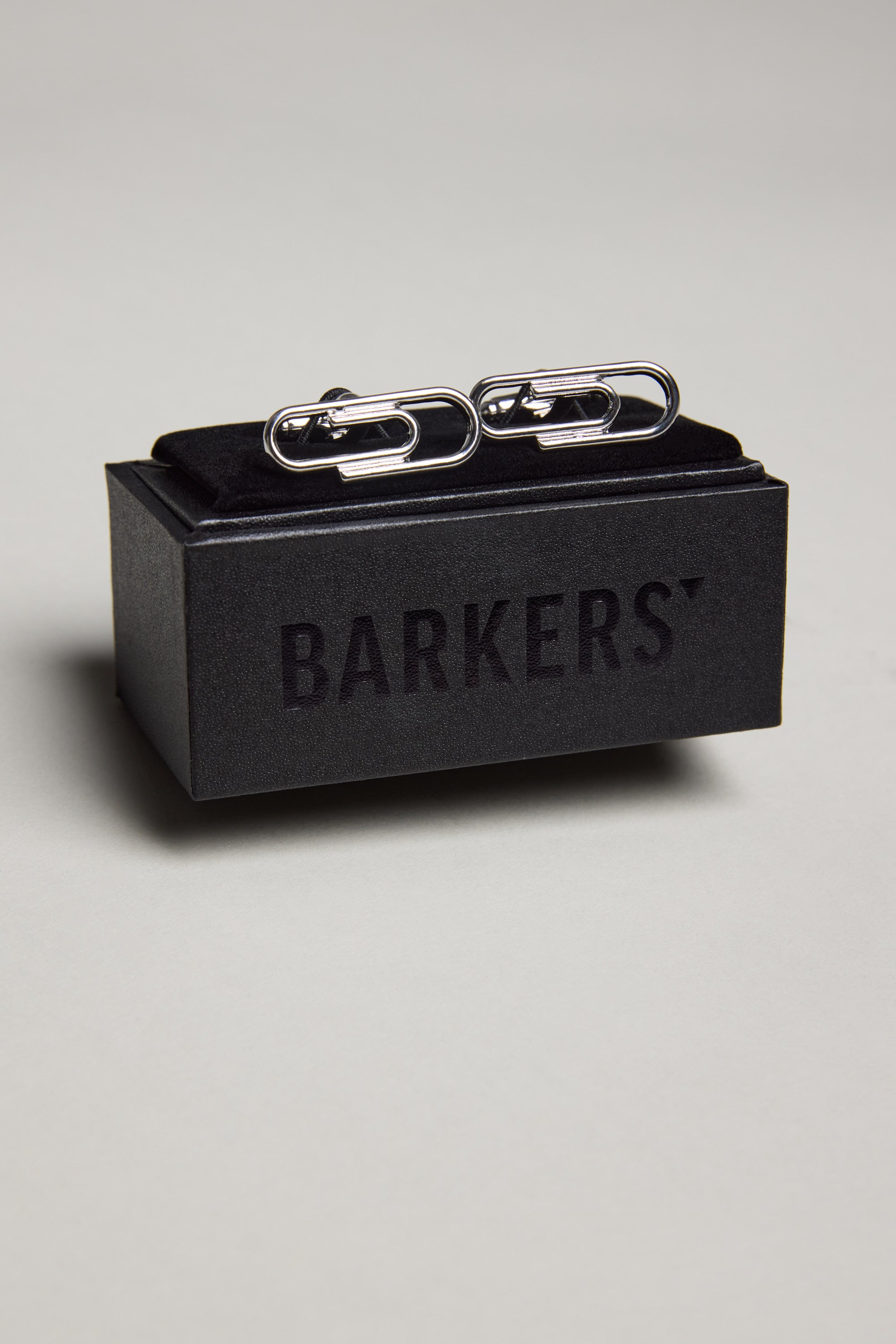 The Barkers Paper Clips Cufflink, featuring a polished silver finish, is displayed on a black box with the Barkers logo—perfect for adding elegance to French cuff shirts. The set rests on a light-colored surface.