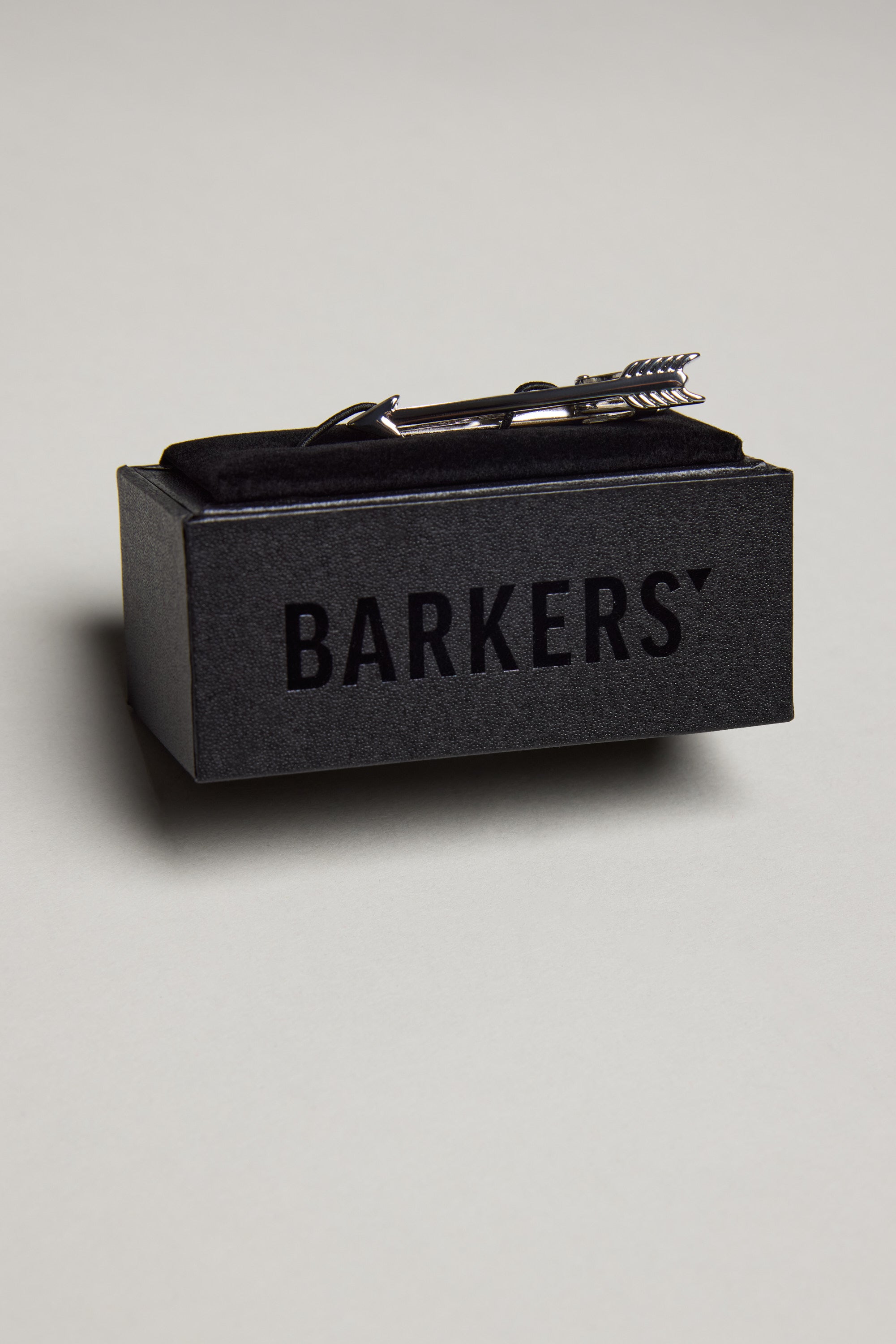 The Barkers Arrow Tie Clip in a polished silver tone sits atop a black box labeled "Barkers," set against a light grey background.