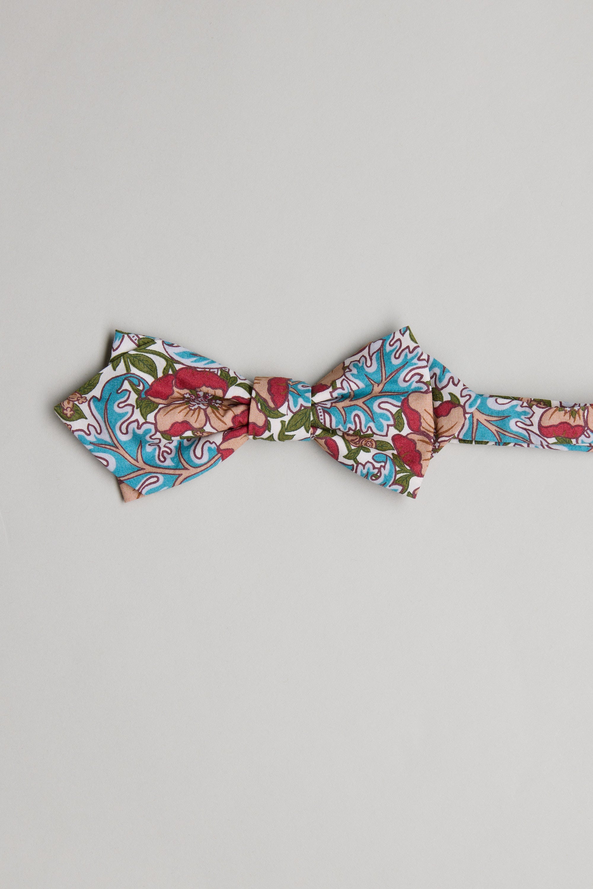 The Liberty Millie Francesca Bow Tie by Liberty is handmade in New Zealand from soft Tana Lawn cotton, featuring a vibrant red, pink, green, and blue floral and leaf pattern on a light grey background.