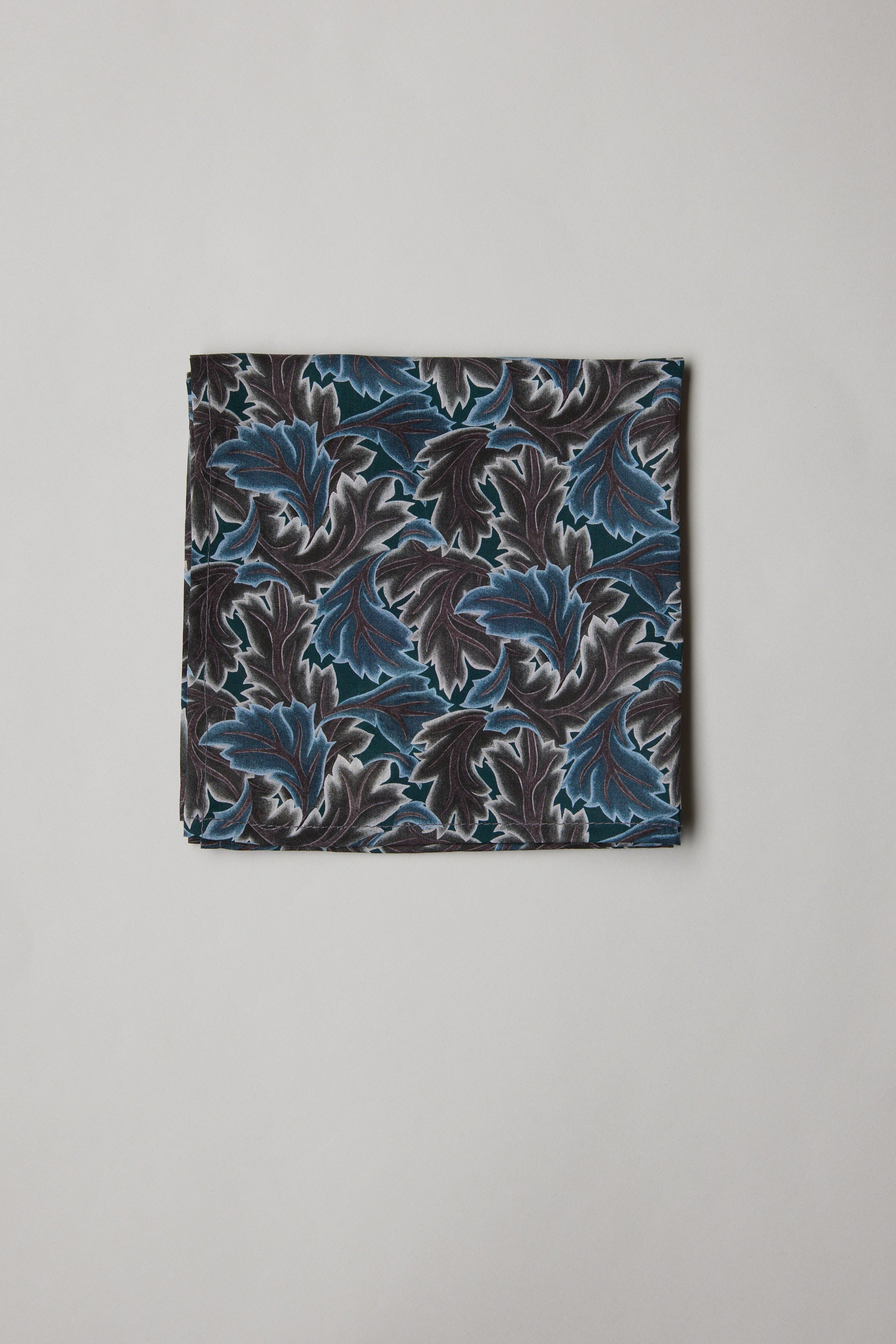 The Liberty Midnight Canopy PS by Liberty is a folded pocket square made from Tana Lawn cotton, showcasing a blue and grey leaf print on a light background.