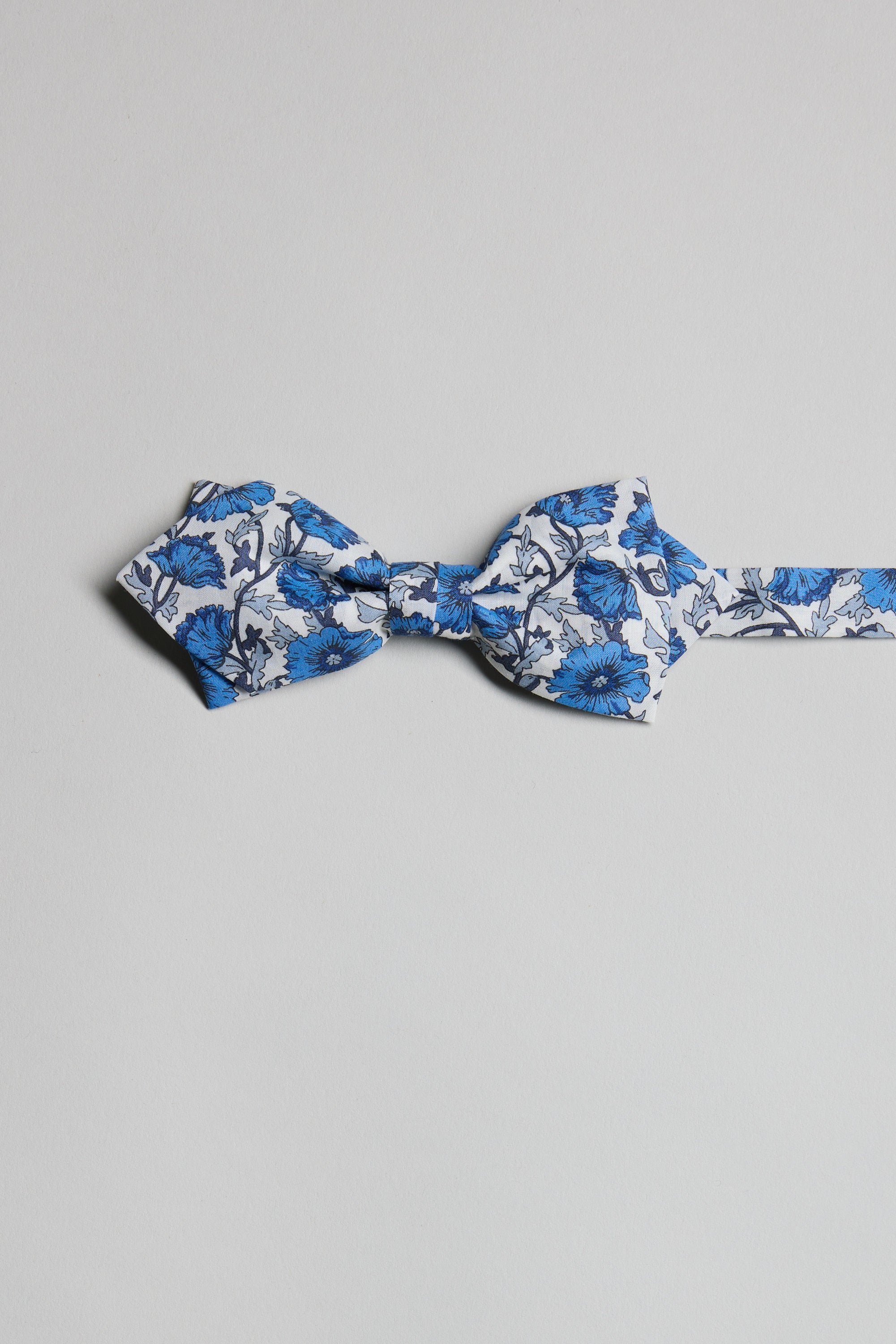 The Liberty Astell Reece Bow Tie by Liberty in a blue and white floral print is laid flat on a plain light gray background, highlighting its timeless elegance.