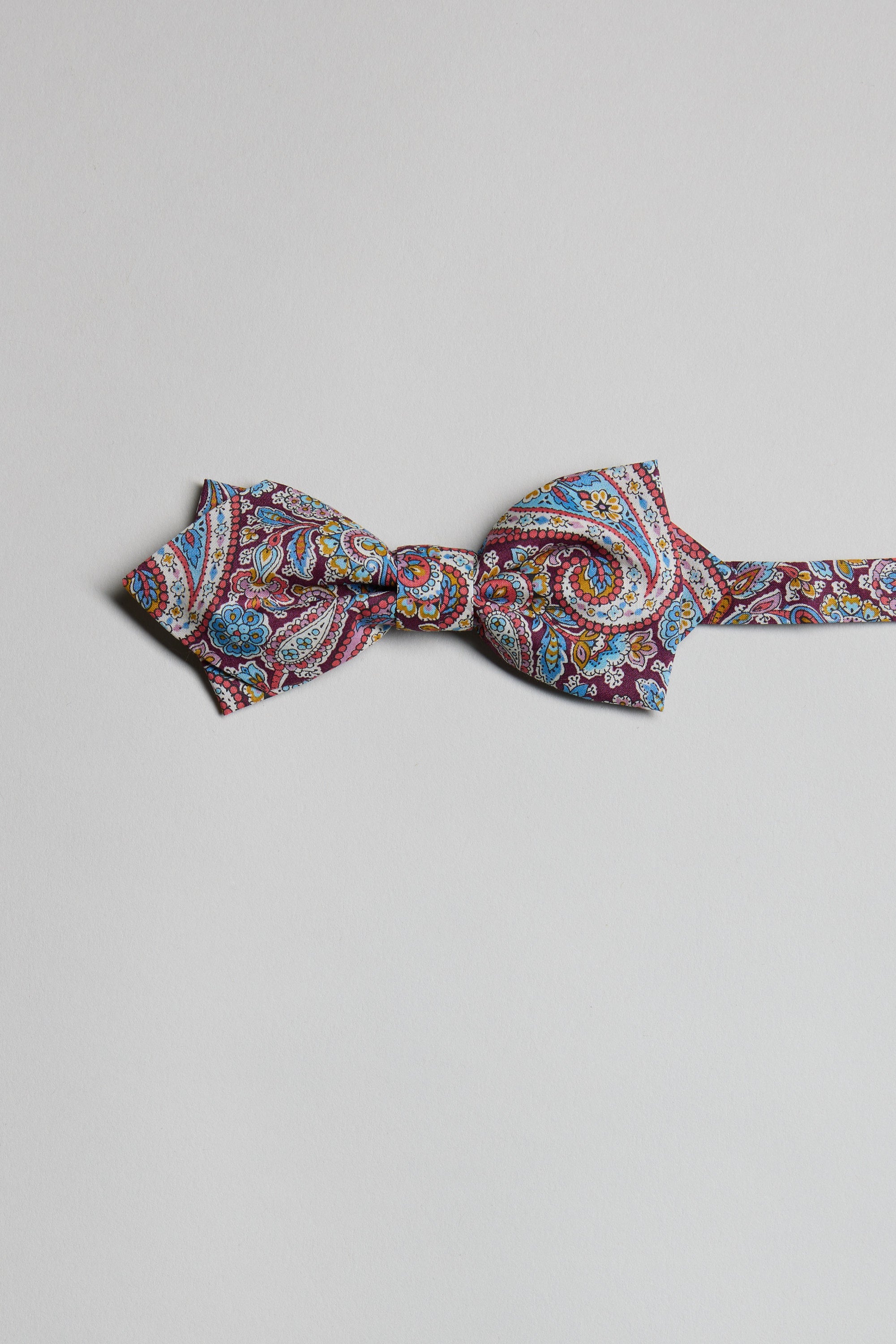 The Liberty Lee Manor Bow Tie by Liberty features a vibrant paisley print in red, blue, yellow, and white tones. Handmade in New Zealand, it is showcased against a plain light gray background.