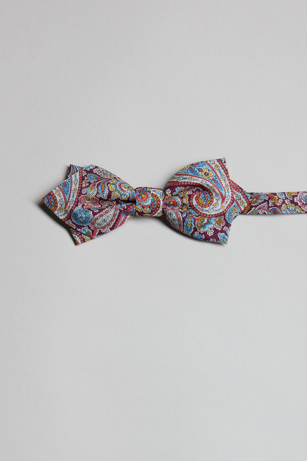 The Liberty Lee Manor Bow Tie by Liberty features a vibrant paisley print in red, blue, yellow, and white tones. Handmade in New Zealand, it is showcased against a plain light gray background.