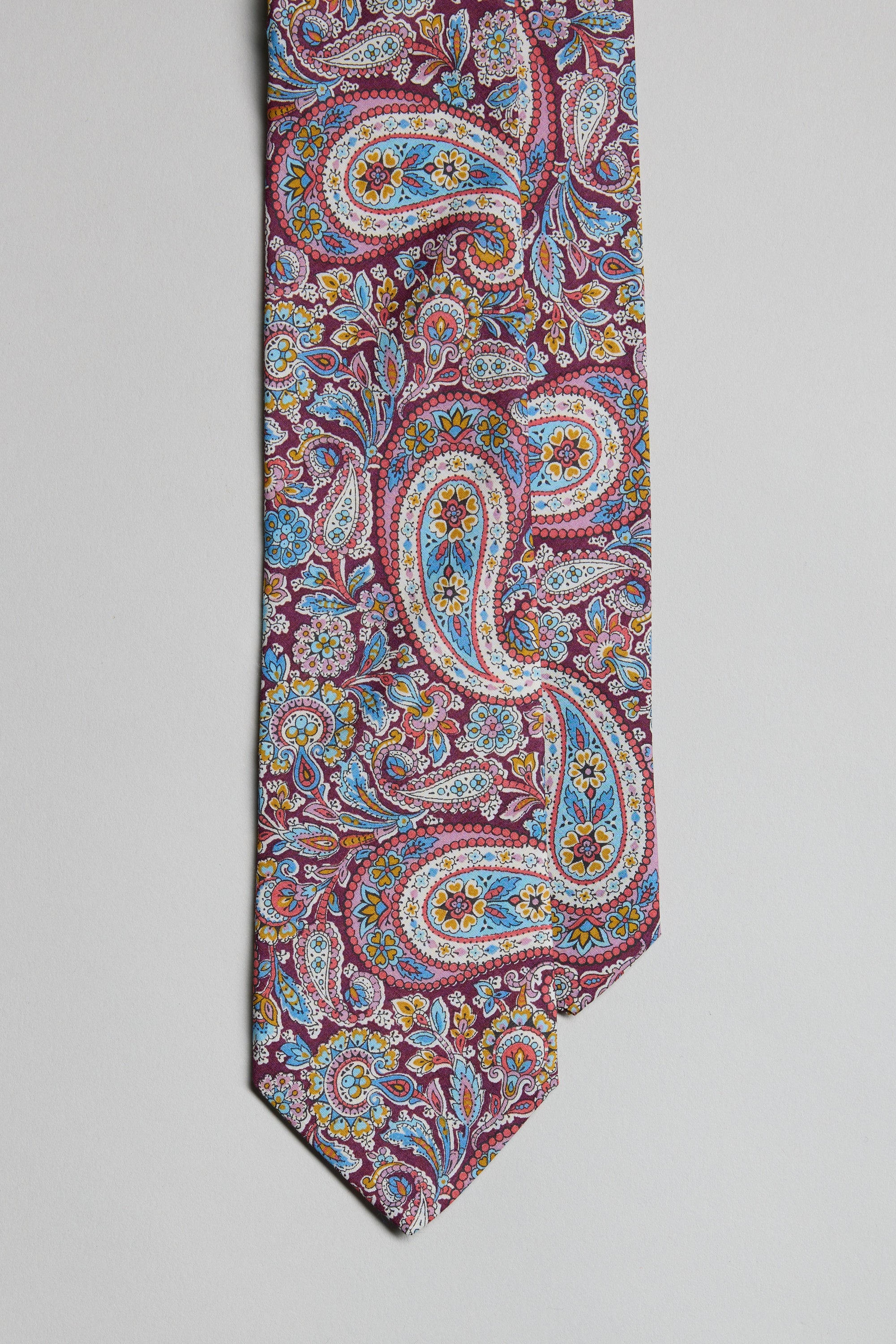 A close-up of the Liberty Lee Manor Tie by Liberty, featuring an intricate paisley pattern in blue, yellow, and white on a reddish-purple background, displayed on a plain light surface.