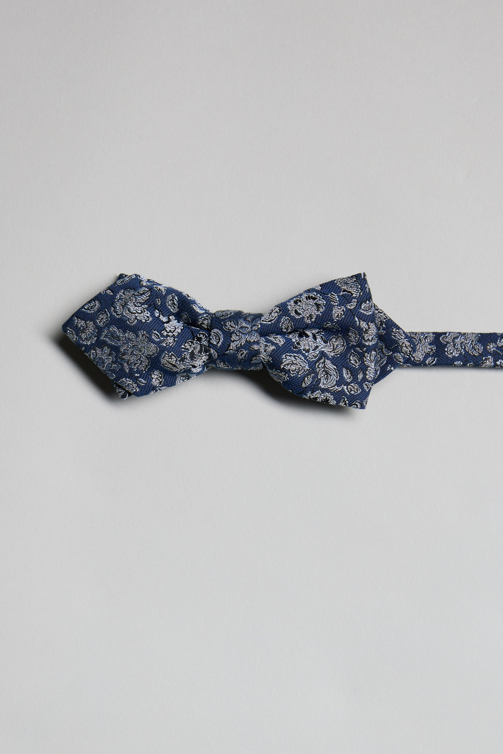 The Parisian Floral Bow Tie by Parisian, featuring a blue paisley pattern, is laid flat on a light gray background with its adjustable neck strap extended to the right.