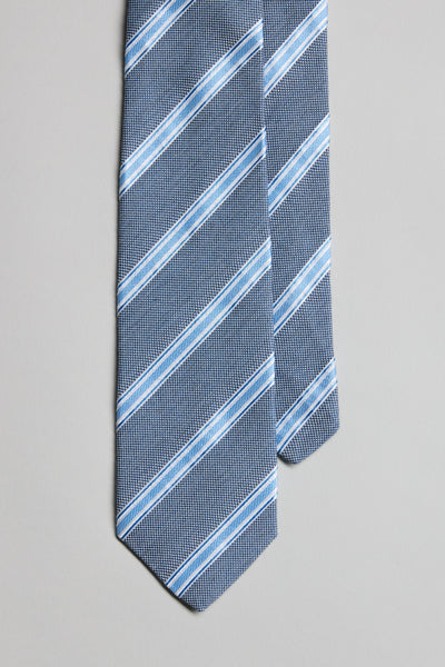 The Parisian Stripe Tie by Parisian, featuring diagonal white and light blue stripes on blue, is handmade in New Zealand and displayed laid flat on a light gray background.