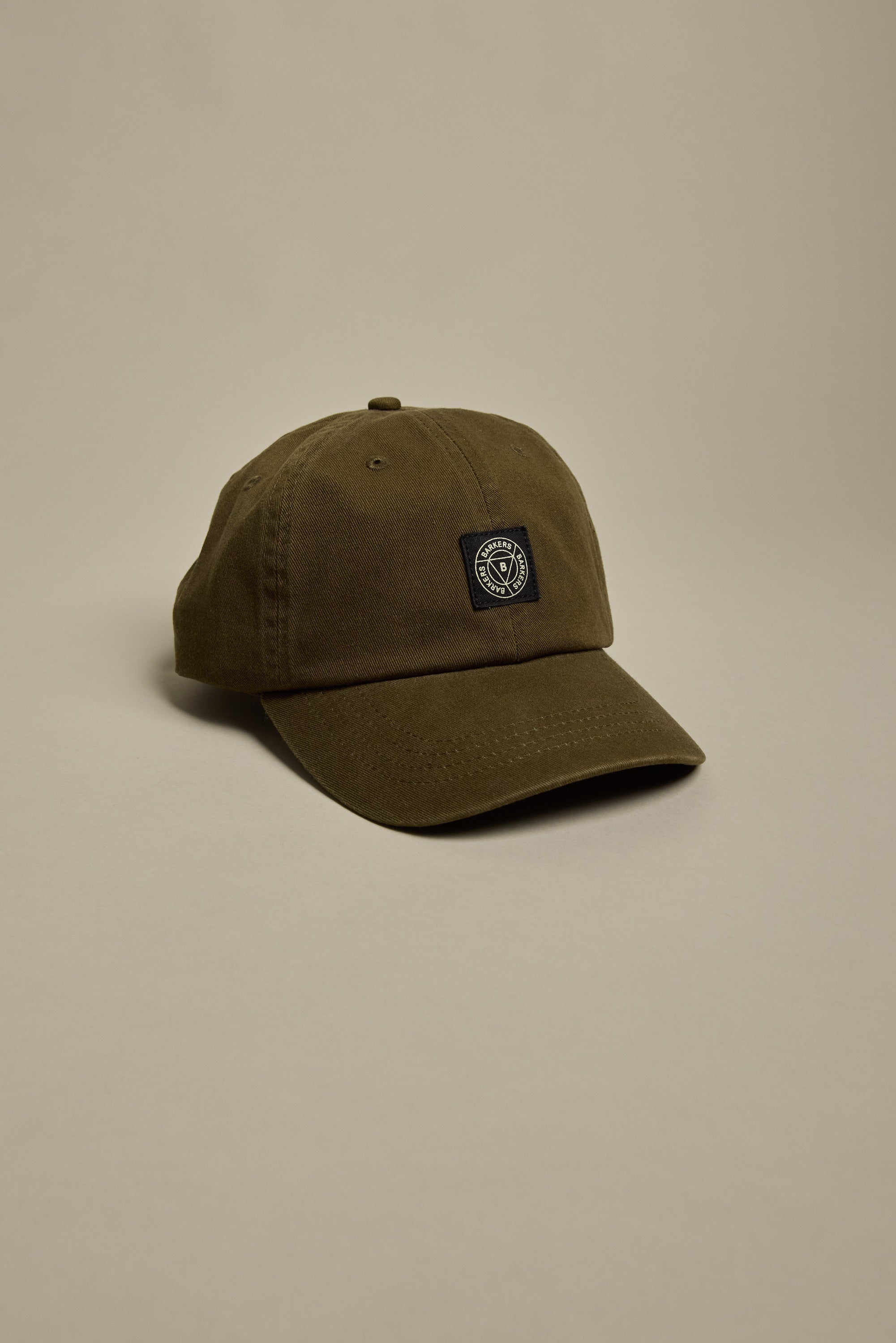 The Barkers Washed Cotton Cap by Barkers features a plain olive-green vintage wash, curved brim, and a small black and white circular geometric embroidered patch on the front, shown against a neutral beige background.