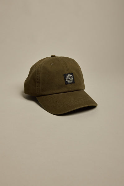The Barkers Washed Cotton Cap by Barkers features a plain olive-green vintage wash, curved brim, and a small black and white circular geometric embroidered patch on the front, shown against a neutral beige background.