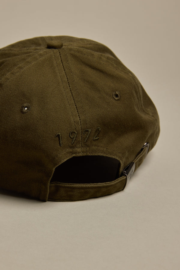 The Barkers Washed Cotton Cap in olive green is shown from the back, with "1972" embroidered above the adjustable strap. The background is a plain light beige.