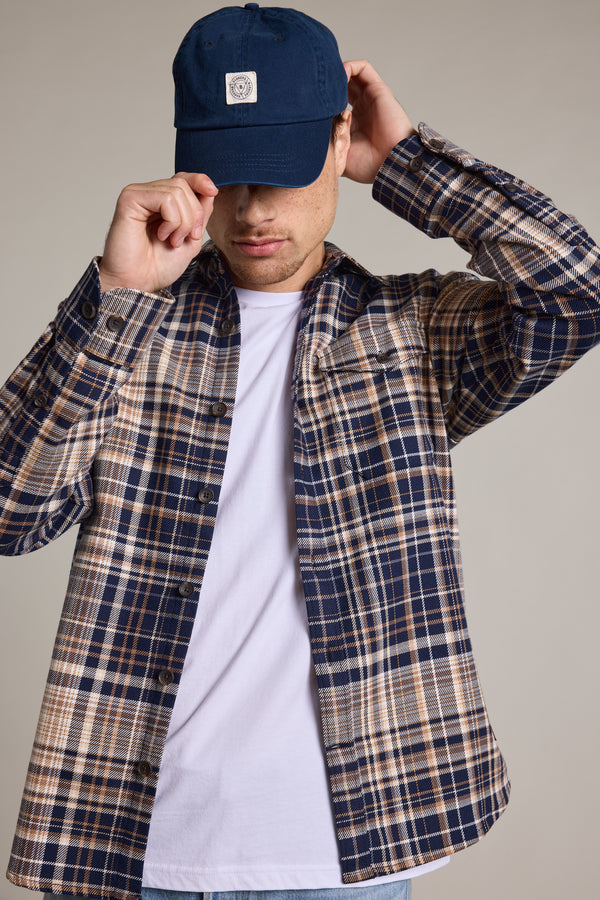 A person wearing a blue plaid shirt, white t-shirt, and a Barkers Washed Cotton Cap stands against a neutral background, tilting the Barkers cap to partially cover their face.