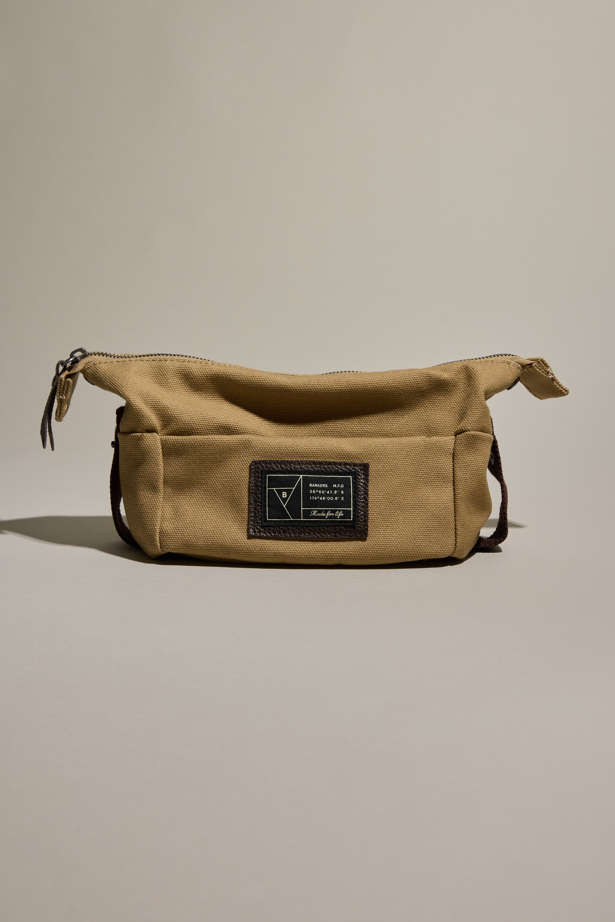 The Barkers Canvas Toiletry Bag by Barkers features a tan canvas design with a zipper closure and a dark rectangular patch with text and graphic on the front, displayed against a neutral background.