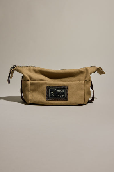 The Barkers Canvas Toiletry Bag by Barkers features a tan canvas design with a zipper closure and a dark rectangular patch with text and graphic on the front, displayed against a neutral background.