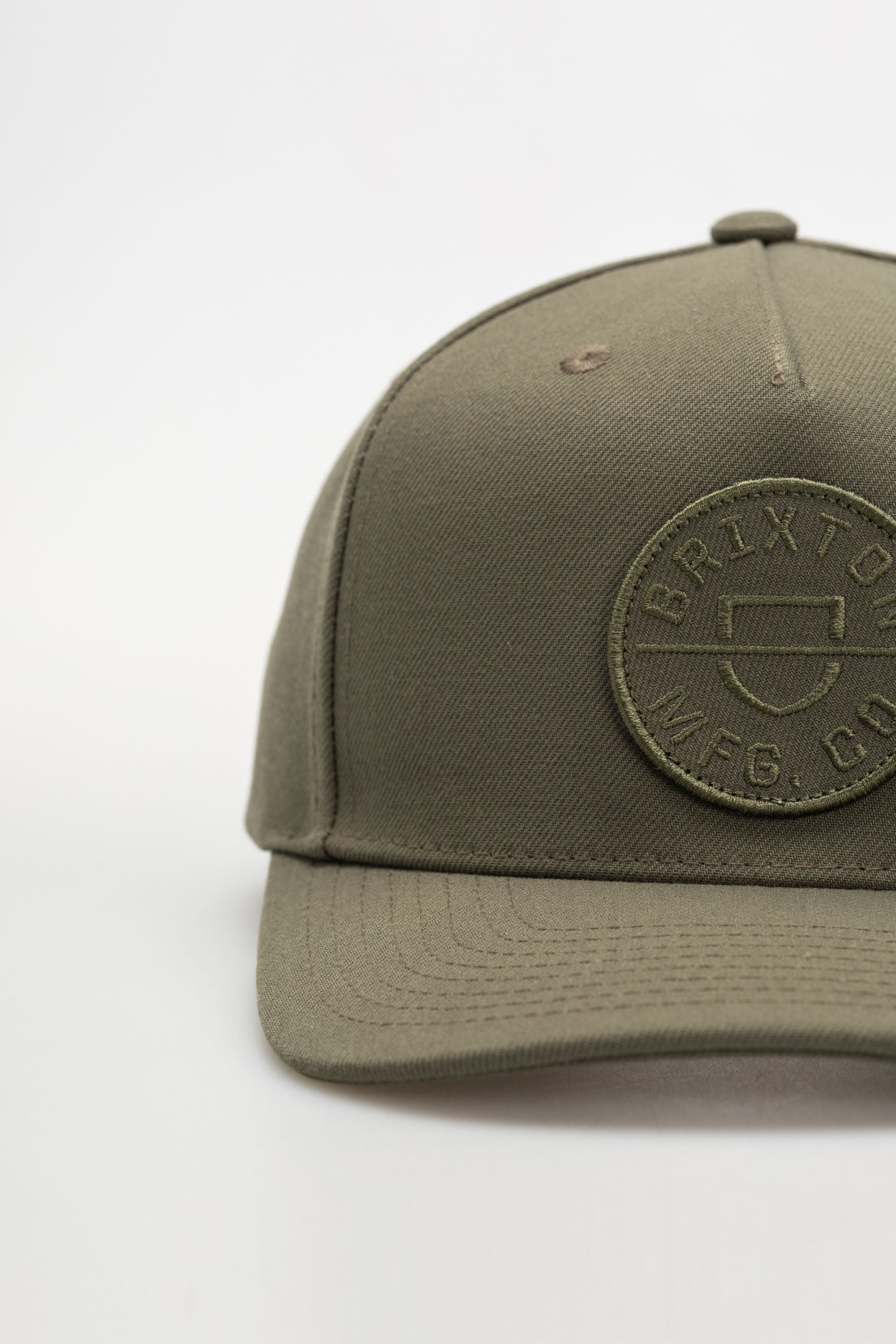 A close-up of the Brixton Crest C MP Snapback, an olive green 5-panel cap with a round embroidered BRIXTON MFG. CO. patch on the front, set against a plain white background.