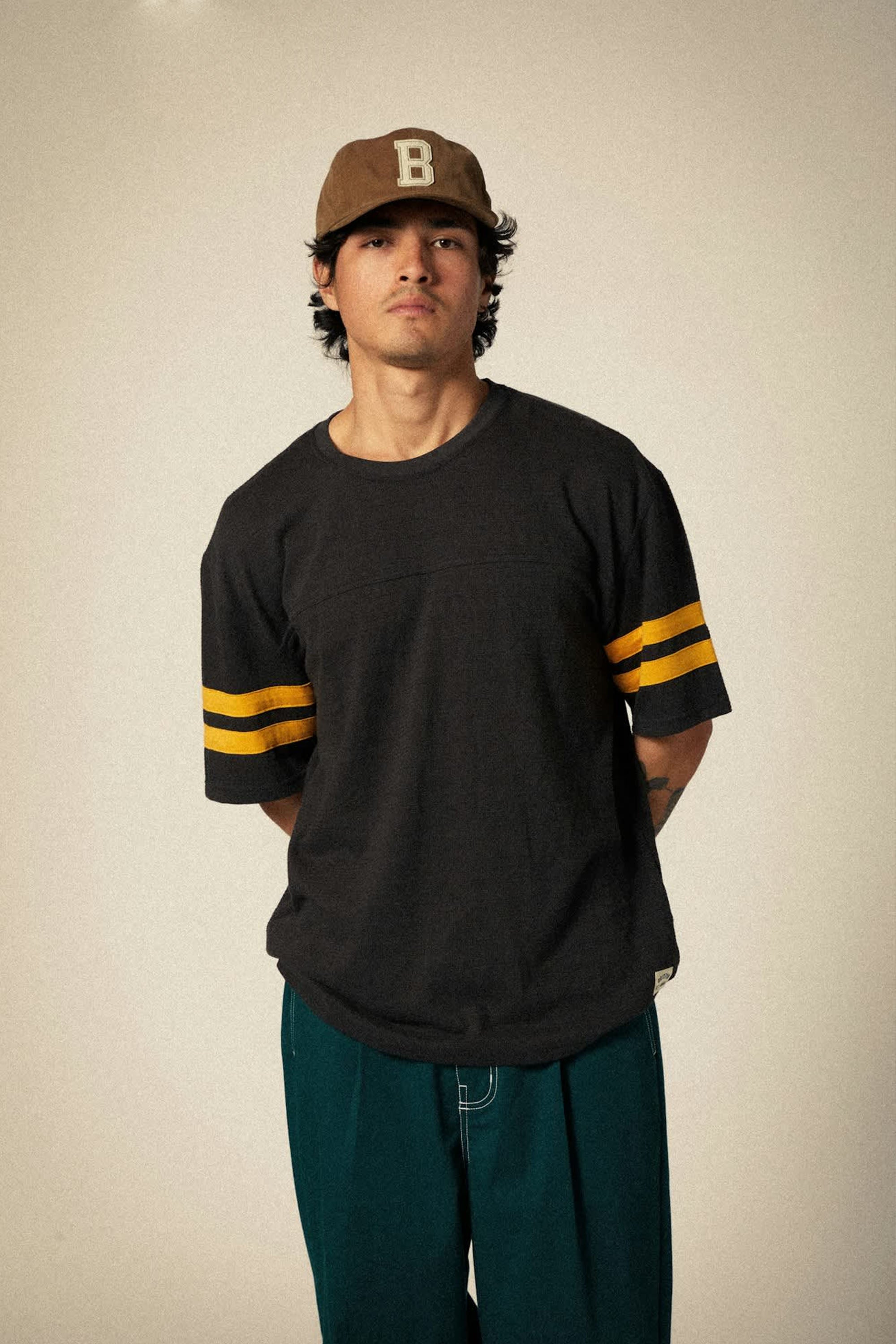 A person wearing the Brixton Big B MP Cap by Brixton, a dark short-sleeve shirt with yellow-striped sleeves, and teal pants stands against a plain background with their hands behind their back.