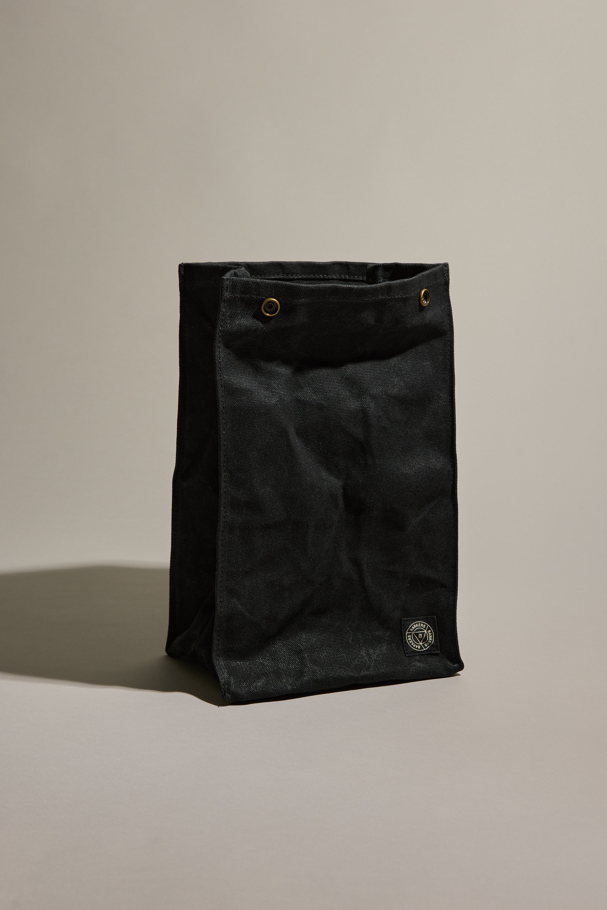 A black Barkers Waxed Lunch Bag stands empty on a beige background, featuring durable cotton canvas, a square base, two metal grommets at the top, and a small circular Barkers logo patch on the lower front.