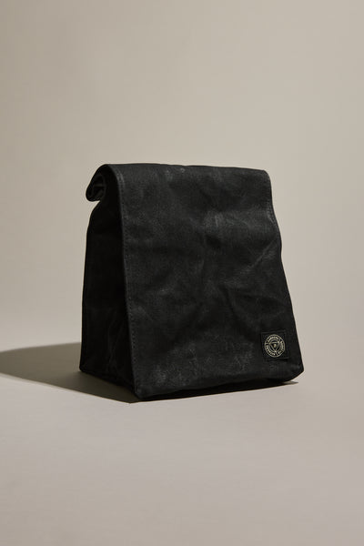 The Barkers Waxed Lunch Bag by Barkers is a black, durable waxed cotton canvas bag with a folded top and a small round logo patch near the bottom right corner, standing upright on a neutral background.