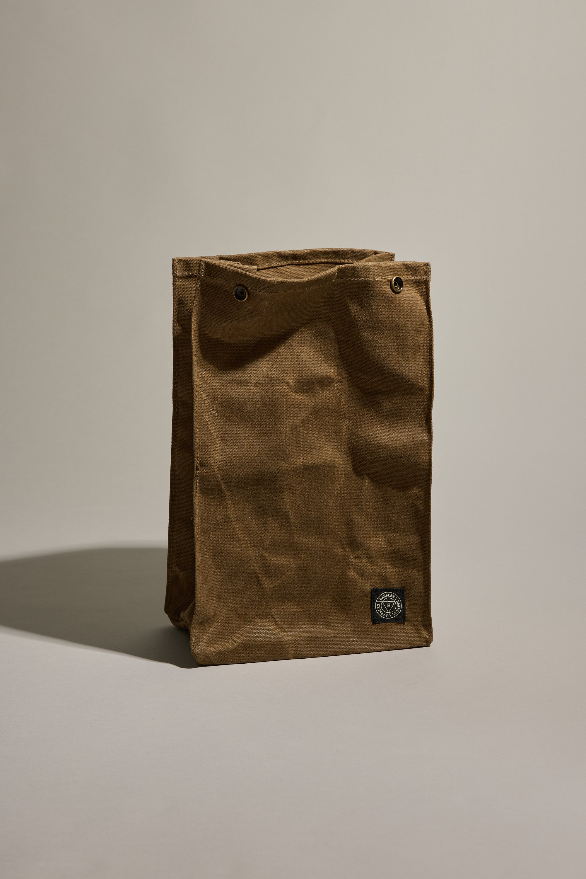 The Barkers Waxed Lunch Bag by Barkers is a brown, rectangular waxed cotton canvas bag with two metal grommets at the top corners and a small black logo patch near the bottom right, shown standing upright on a neutral background.