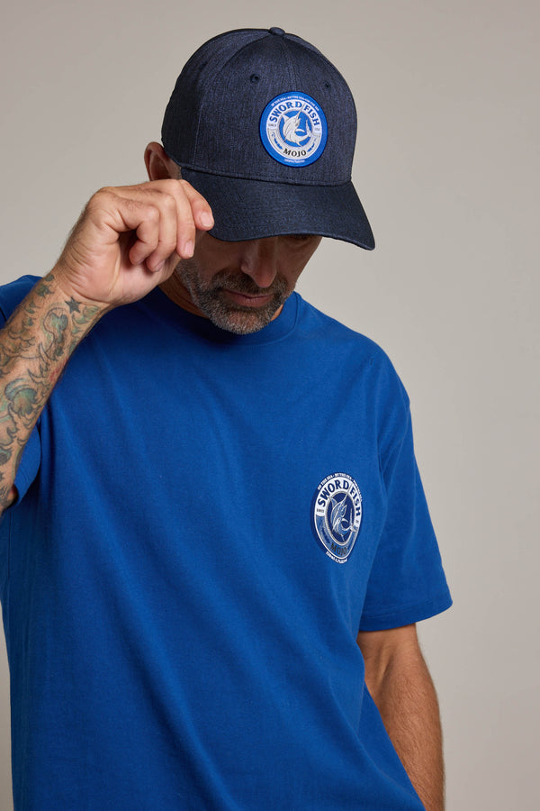 A man in a blue T-shirt wears a Swordfish Mojo Trucker Cap, tilting it down to partially cover his face. His right arm shows visible tattoos.