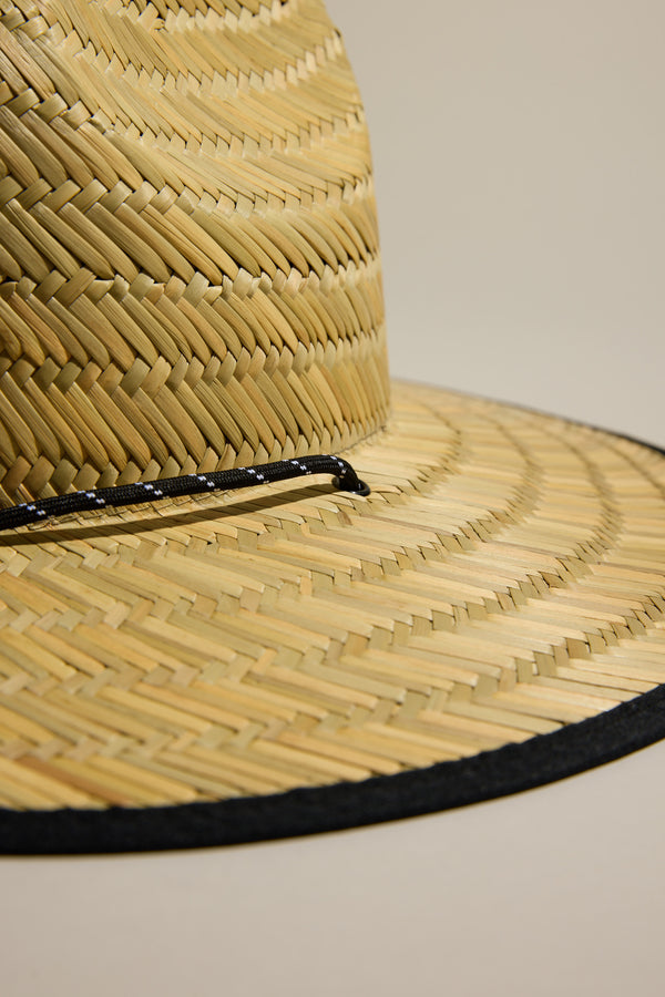 Close-up of the Barkers Straw Hat by Barkers, a summer essential featuring a wide brim, black band, and decorative string above the brim, providing stylish sun protection against a plain beige background.