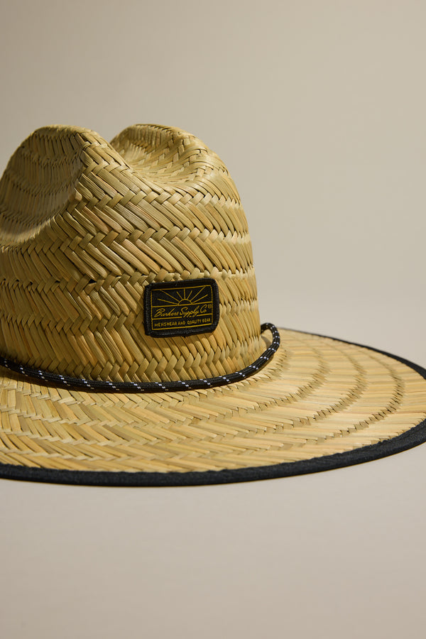 The Barkers Straw Hat by Barkers features a wide brim, black trim, woven pattern, and a rectangular black-and-gold front label. This summer essential provides stylish sun protection against neutral backgrounds.