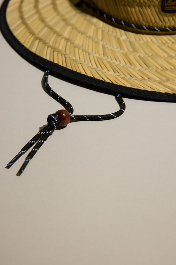 A close-up of the Barkers Straw Hat by Barkers, featuring a wide brim and black chin cord with wooden bead, rests on a light beige surface. Part of the brim and most of the cord are visible—a perfect summer essential for sun protection.