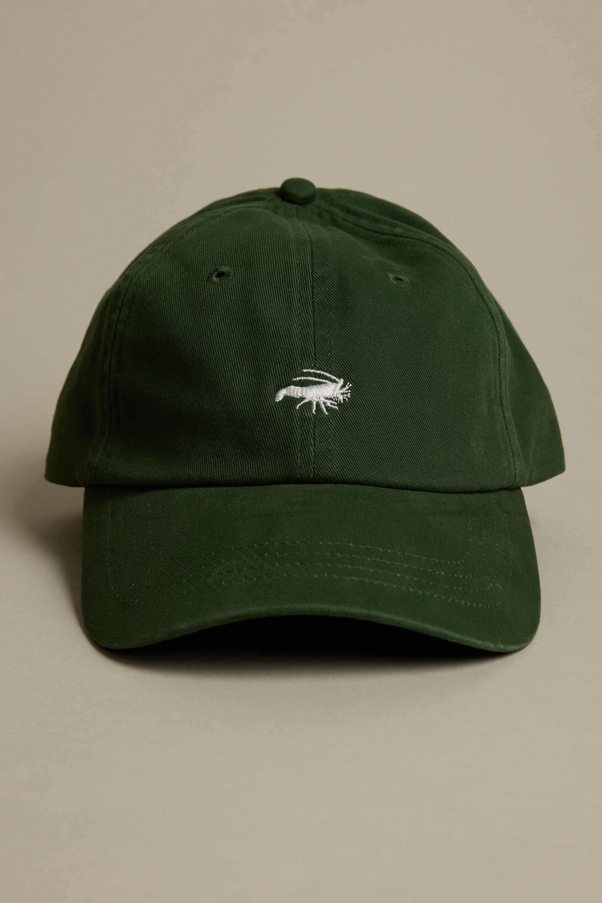 The Barkers Icon Cotton Cap is a dark green adjustable cotton cap with a small white embroidered fish, inspired by the Barkers 1972 logo, photographed on a plain beige background.