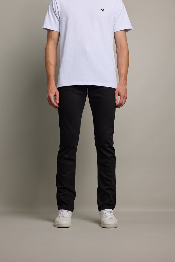 A person stands facing forward against a plain background, wearing a white short-sleeve T-shirt, Barkers Rambler Denim Slim Jean, and white sneakers. Only the body from shoulders down is visible.