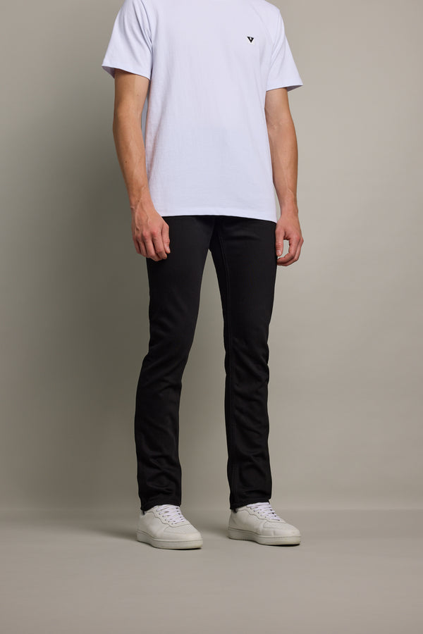 A person stands against a plain background, wearing a white short-sleeve T-shirt, Barkers Rambler Denim Slim Jean in black sustainable denim, and white sneakers. The person's head is not visible in the image.