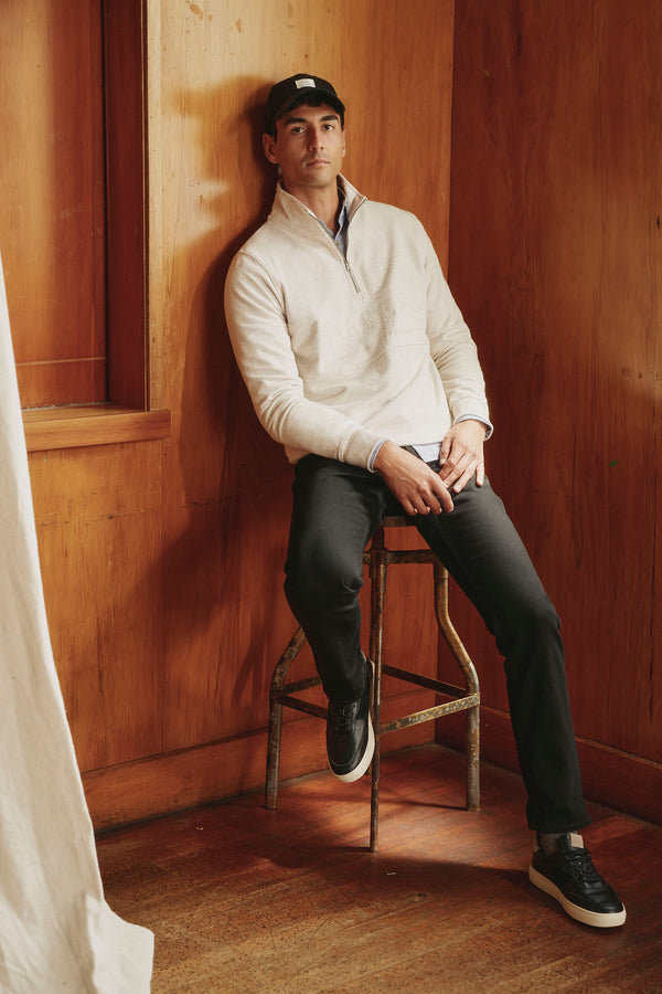 A man in a white pullover, black cap, and black sneakers sits on a stool by a wood-paneled wall wearing Barkers Wanderer Denim Straight Jean. He looks relaxed at the camera as light enters from the left.