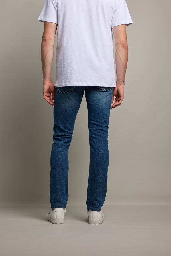 A person stands facing away from the camera against a neutral background, wearing Barkers Rambler Denim Slim Jean, a white t-shirt, and white sneakers.