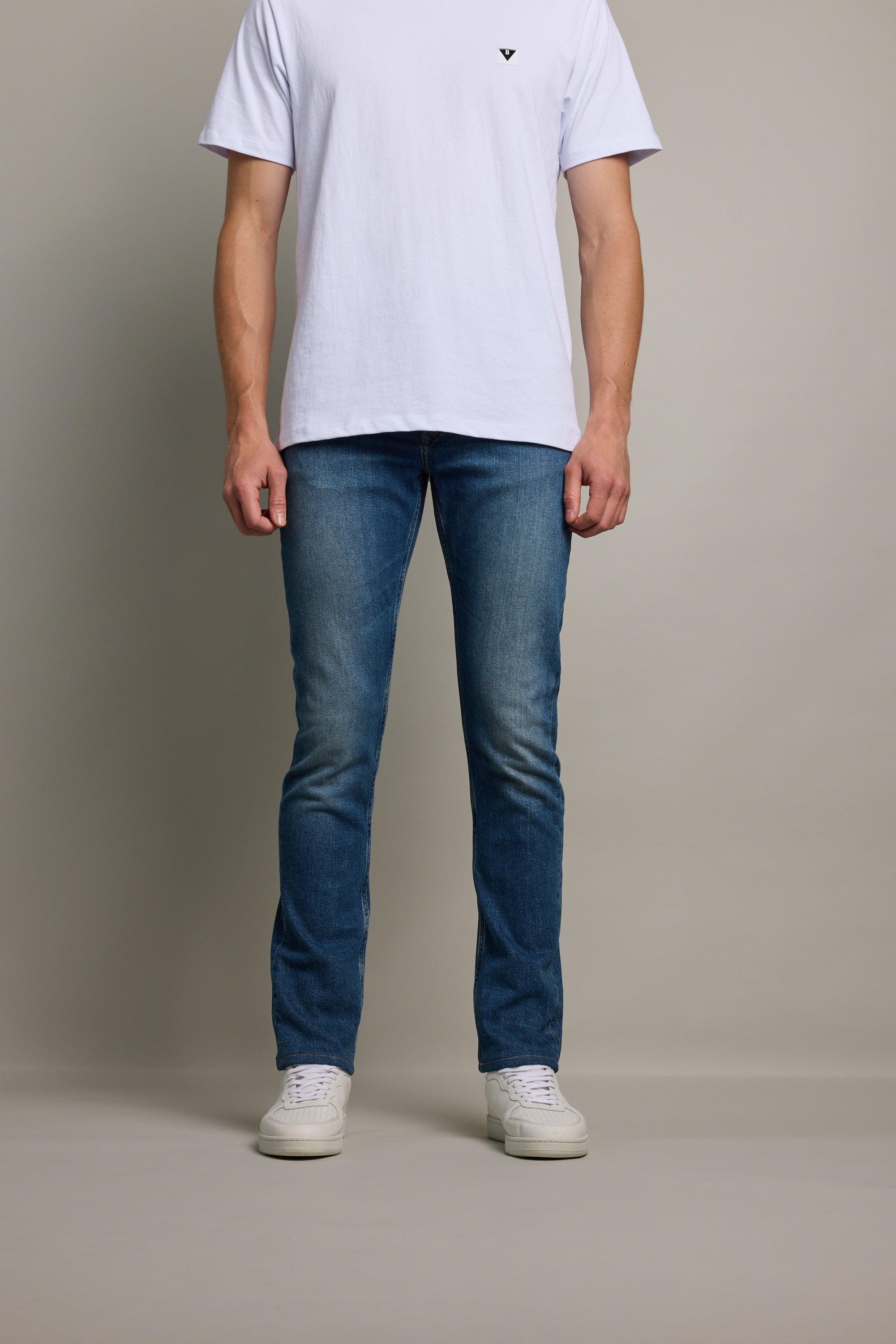 A person stands against a plain background, wearing Barkers Rambler Denim Slim Jeans, a white t-shirt, and white sneakers. The image is cropped at the shoulders to highlight the jeans and lower body.