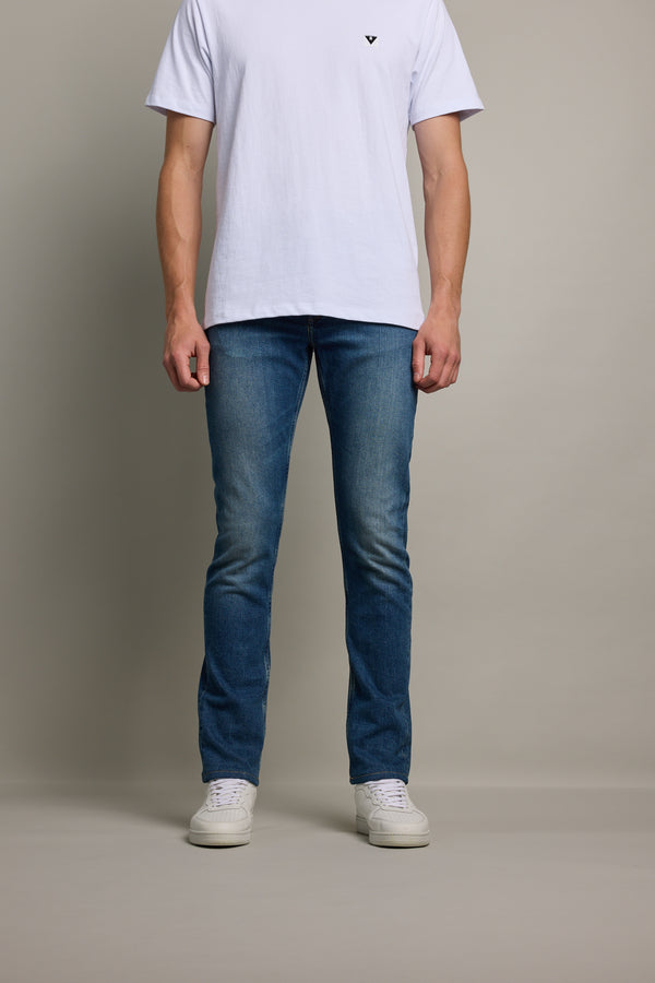 A person stands against a plain background, wearing Barkers Rambler Denim Slim Jeans, a white t-shirt, and white sneakers. The image is cropped at the shoulders to highlight the jeans and lower body.