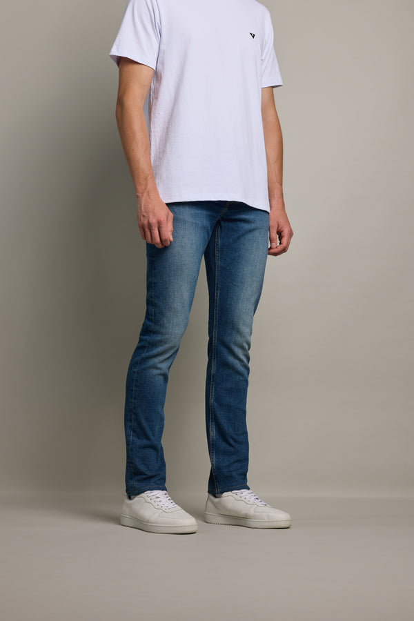 A person stands against a plain background, wearing a white short-sleeve t-shirt, white sneakers, and Barkers Rambler Denim Slim Jean in classic blue. Their head is not visible in the image.