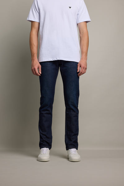 A person stands against a plain background wearing a white t-shirt, Barkers Rambler Denim Slim Jean, and white sneakers. The photo is cropped at the shoulders, showing only the torso and legs.