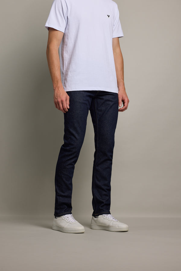 A man stands against a plain background, wearing Barkers Rambler Denim Slim Jeans, a white short-sleeve t-shirt, and white sneakers. His face is not visible in the image.