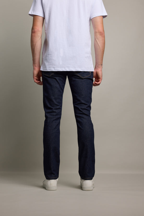 A person stands facing away from the camera, wearing a plain white T-shirt, Barkers Rambler Denim Slim Jean made from sustainable denim, and white sneakers against a neutral background.