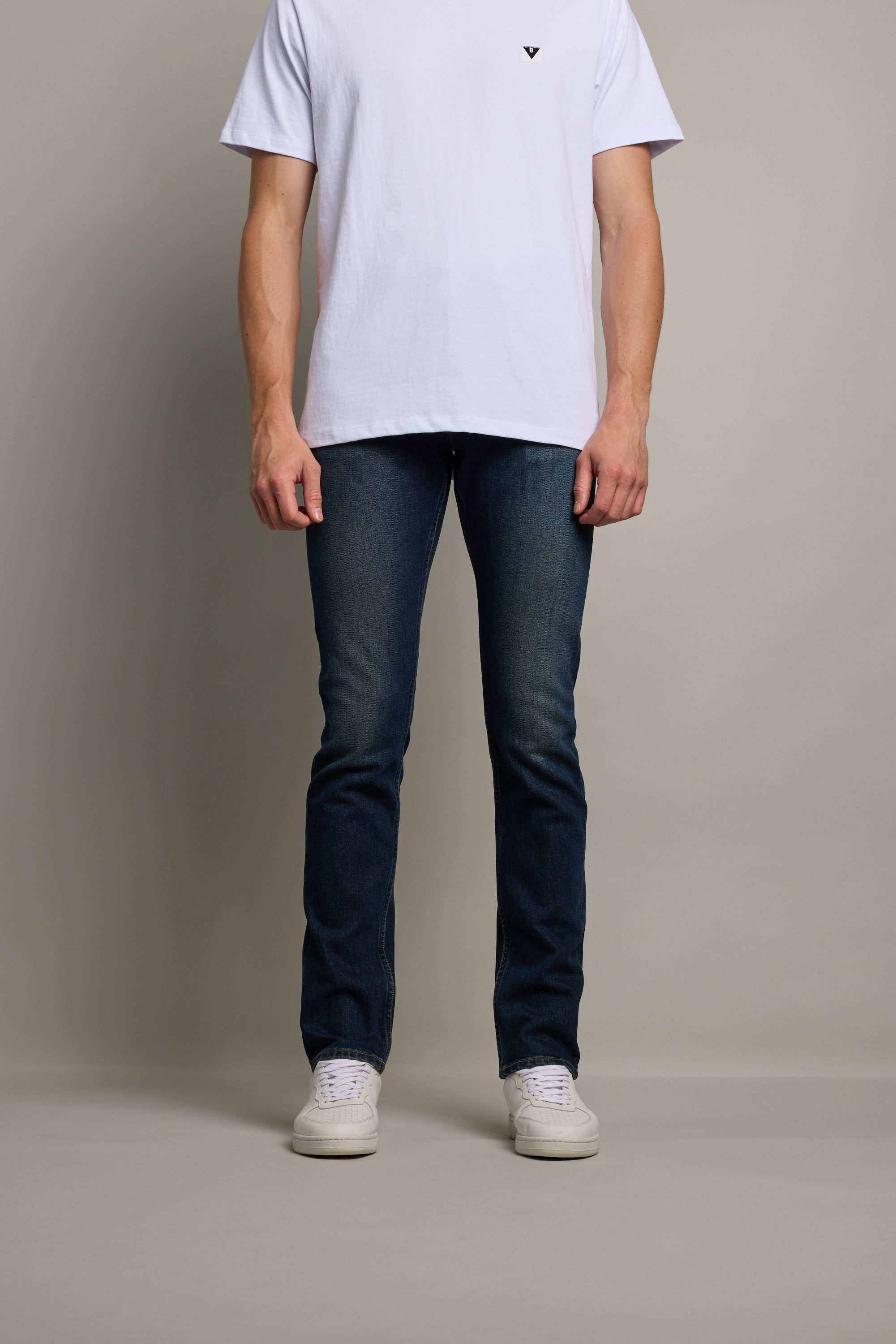 A person wearing a plain white t-shirt, Barkers Rambler Denim Slim Jean in dark blue, and white sneakers stands against a light background. The image does not show the person's head.