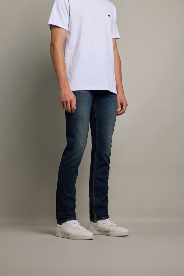 A person stands against a plain background wearing a white short-sleeve t-shirt, Barkers Rambler Denim Slim Jean in dark blue, and white sneakers. The person's face is not visible.