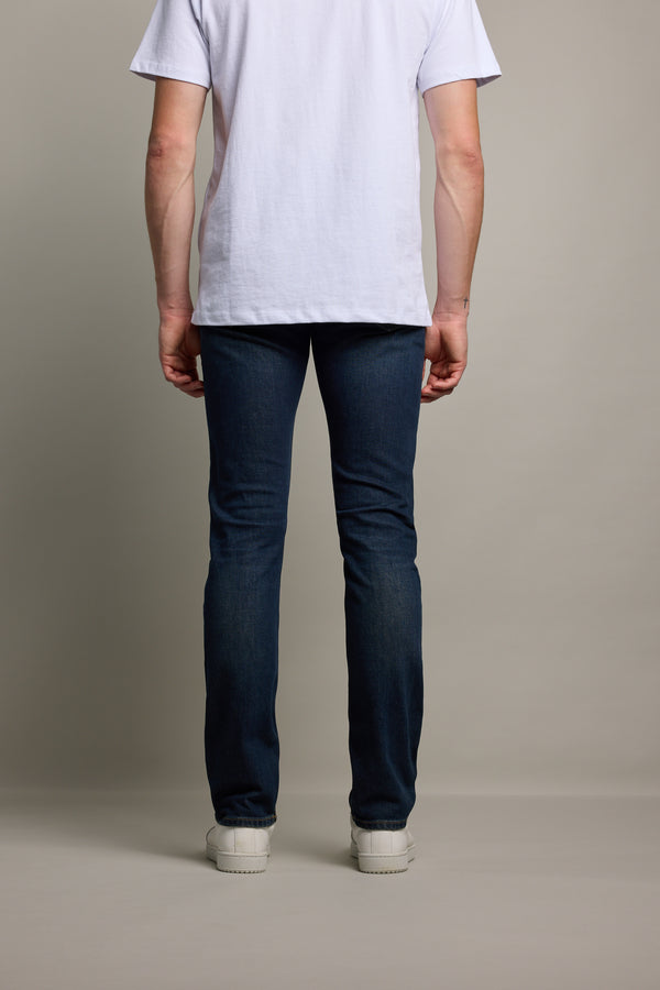 A person stands facing away from the camera, wearing a white short-sleeve t-shirt, Barkers Rambler Denim Slim Jean in dark blue, and white sneakers against a plain light-colored background.