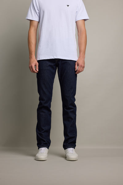 A person, head not visible, stands against a plain background wearing a white short-sleeve T-shirt, Barkers Nomad Denim Taper Jean in dark blue, and white sneakers.