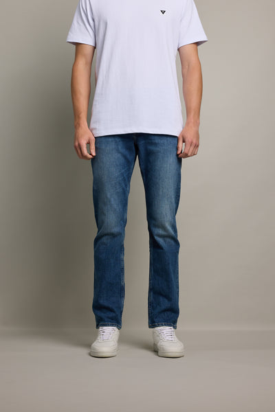 A person stands against a plain background, visible from shoulders down, arms at their sides, wearing a white short-sleeve t-shirt, Barkers Wanderer Denim Straight Jean in sustainable denim, and white sneakers.