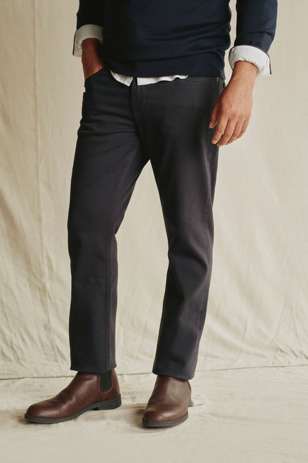 Against a neutral background, a person’s lower body and hand are visible, wearing Barkers’ Wanderer Straight Coloured Jean made from sustainable denim, paired with dark pants, brown Chelsea boots, a navy sweater, and white shirt.