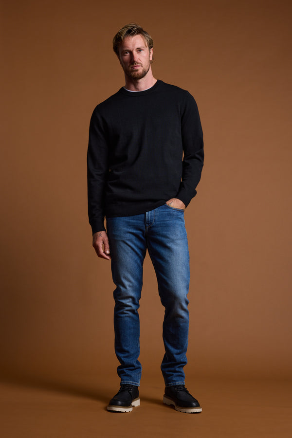 A man with short blond hair and light stubble stands against a brown background wearing the Barkers Skyland ZQrx Merino Crew, blue jeans, and black shoes, one hand in his pocket as he looks at the camera.