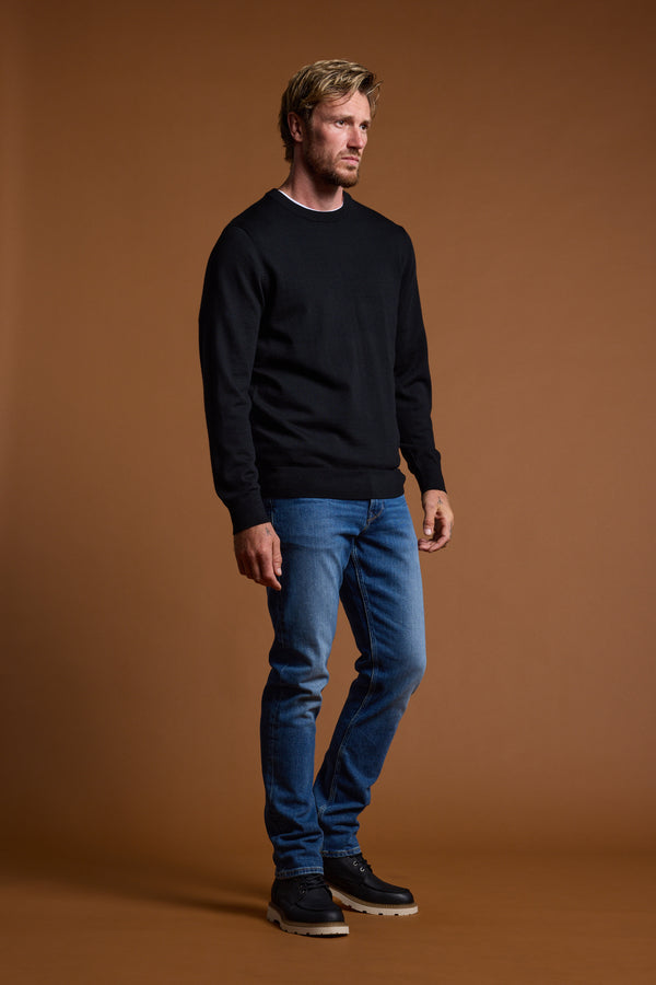 A man with short blond hair and a beard stands against a plain brown background, wearing the Barkers Skyland ZQrx Merino Crew sweater, blue jeans, and black shoes. He gazes off to the side with a neutral expression.