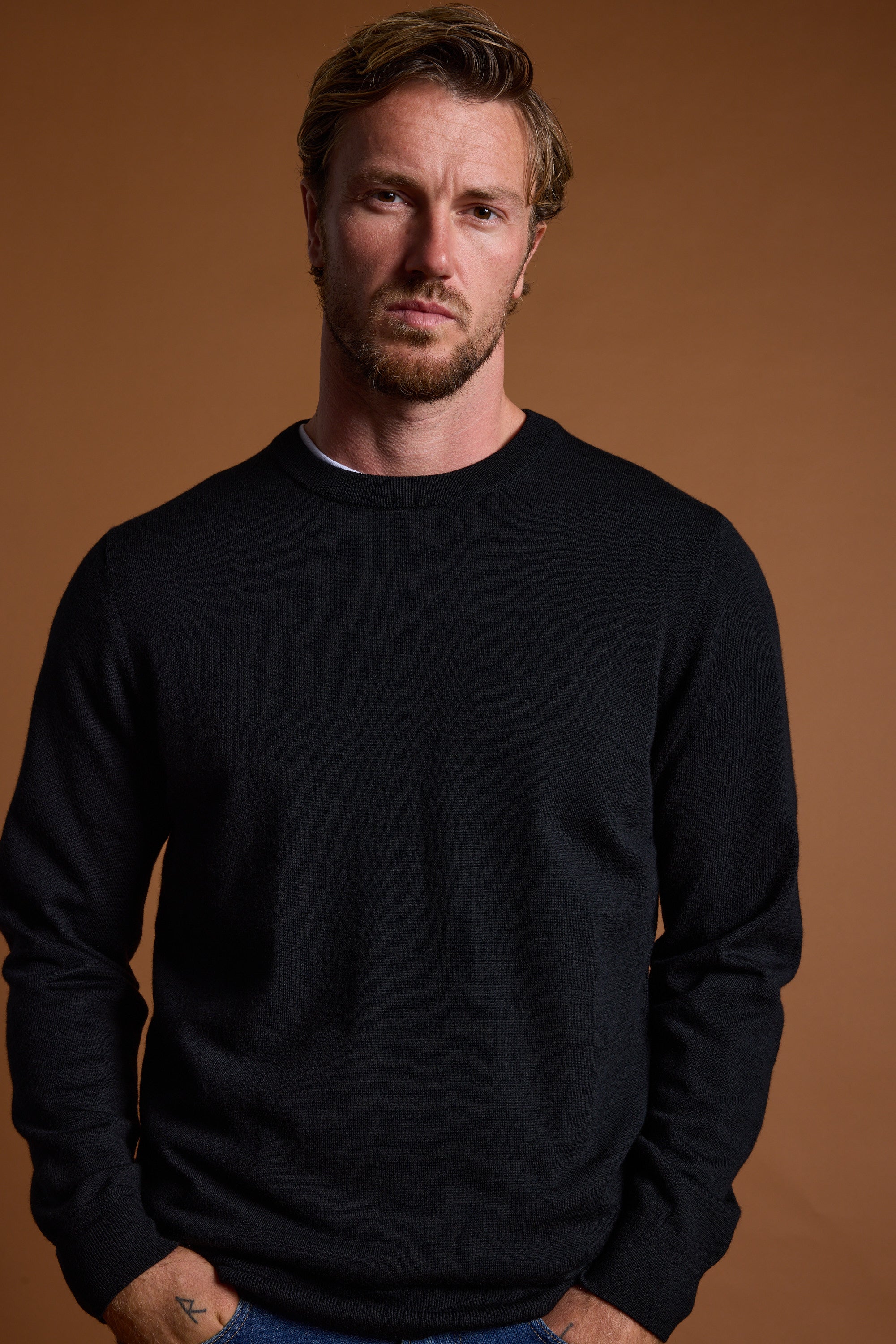 A man with short light brown hair and a beard wears the Barkers Skyland ZQrx Merino Crew, made from ethically sourced wool, standing against a plain brown background with his hands in his pockets, looking at the camera.